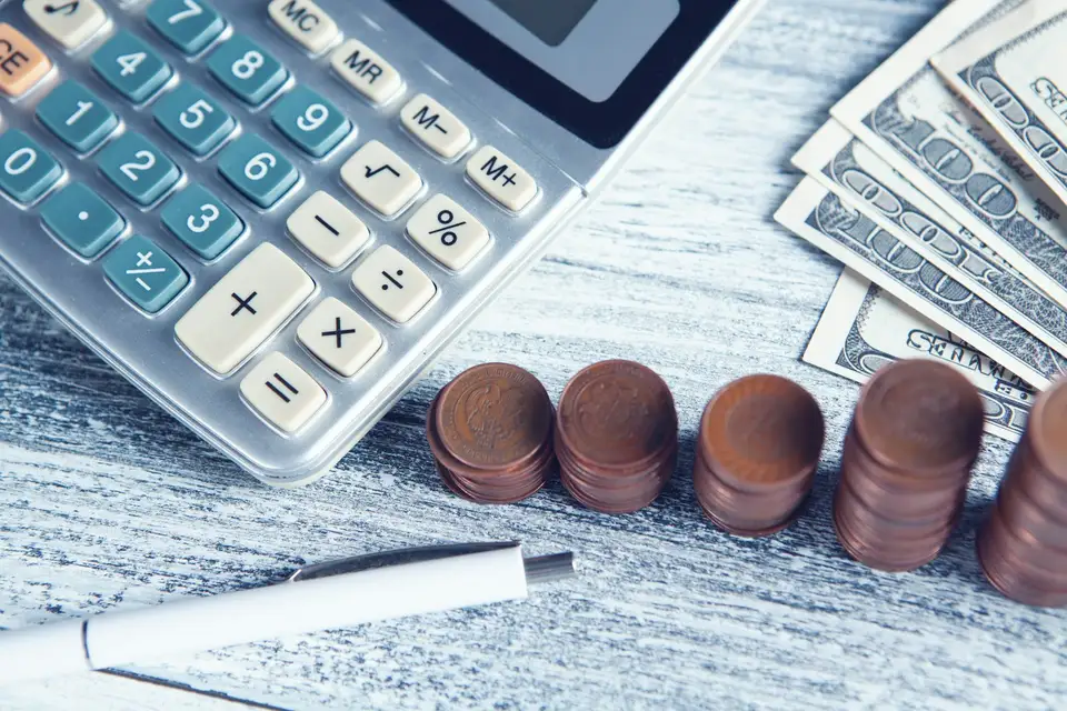 Calculadora, un esfero, billetes de dólar y pilas de monedas sobre una mesa de madera clara