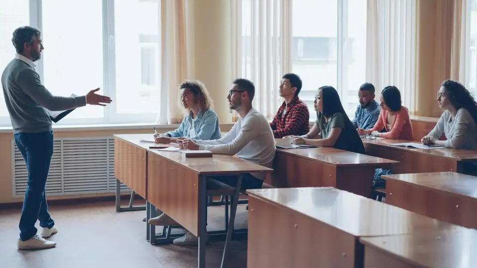 Instructor de pie hablando frente a un grupo de adultos sentados en pupitres, en un aula