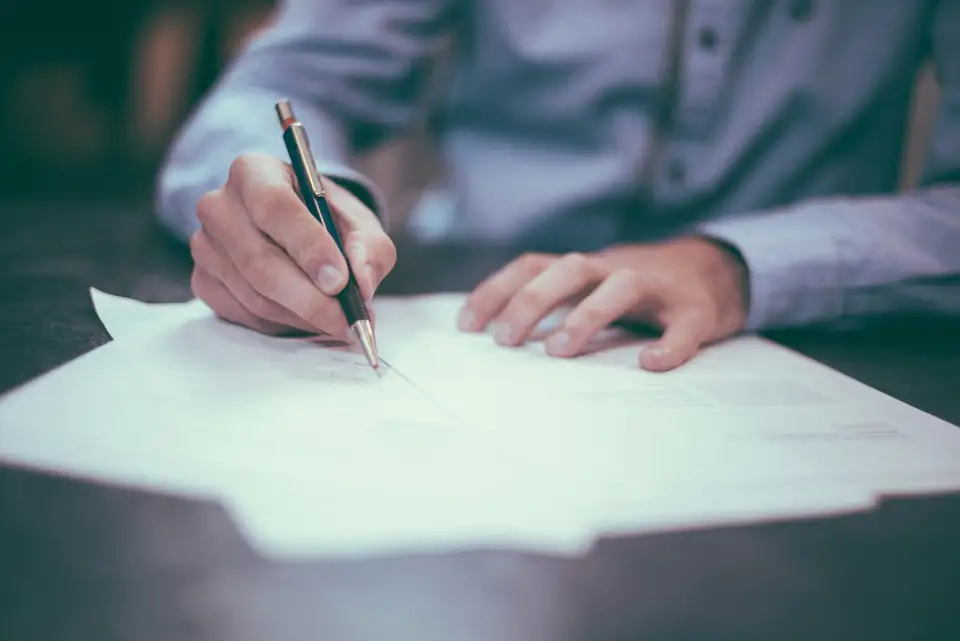 Mano firmando un documento con un esfero, sobre un escritorio oscuro